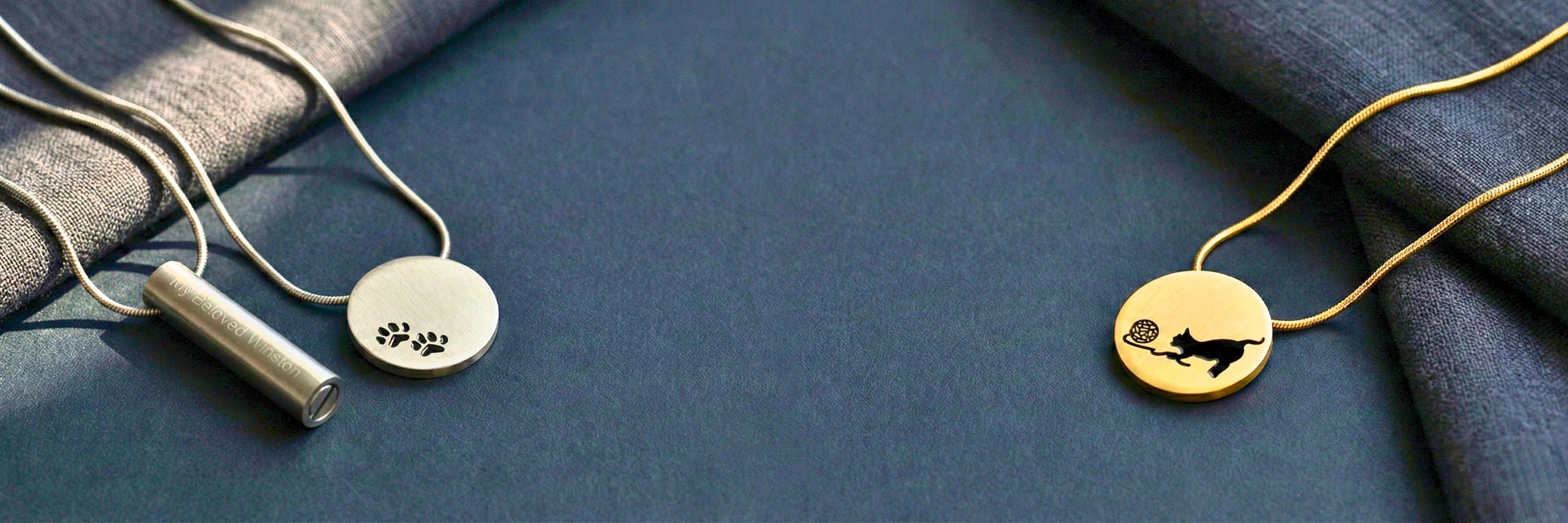 Pet memorial jewelry collection featuring silver paw print pendant with cylinder urn and gold cat silhouette necklace on dark blue linen — Magnolia Memorial Marketplace